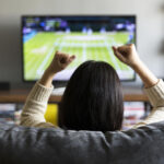 Young woman enthusiastically watching a tennis match on live sports streaming service on television, cheering with excitement.