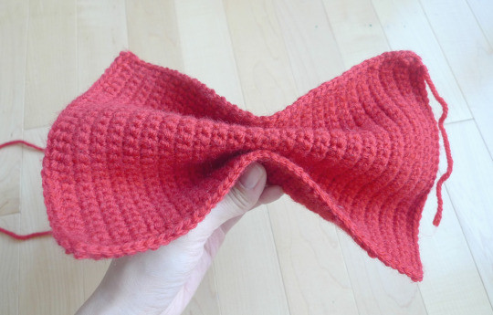 Hands pinching the red crocheted rectangle in the center to demonstrate how to shape it into a bow.