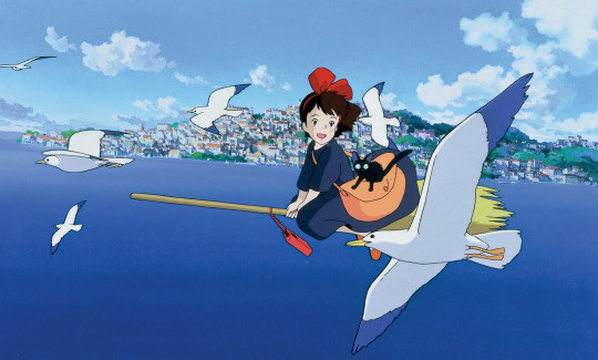 Cosplayer dressed as Kiki from Kiki's Delivery Service, wearing a large red bow, black dress, and holding a brown satchel.