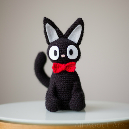 Close up of a handmade Jiji amigurumi, a small black cat with red eyes, sitting and looking cute, promoting the Jiji crochet pattern.