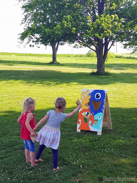 Two children in a park are tossing bean bags into a sandwich board painted to look like a monster mouth.