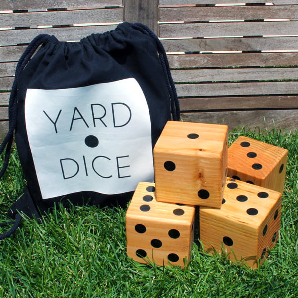 Four large wooden dice with black dots are resting on green grass next to a black and white bag that says, “Yard Dice.”