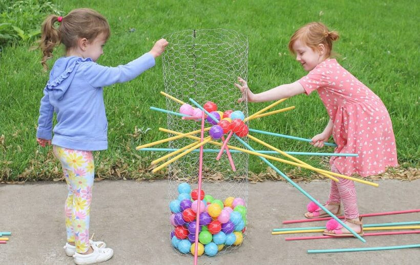 A colorful Ker-Plunk game is set up in a grassy yard and played by two children; the colorful balls are in a wire net, and the pins are in place. 
