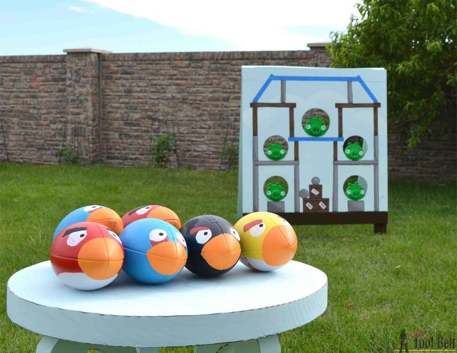 A backyard is shown with a white table; on the table are six footballs that look like Angry Birds and in the background is wooden house cut-out with holes for the footballs. 