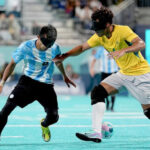 A man in a yellow Brazilian football jersey wearing a black eyeshade plays a football with his foot, while a man in a blue and white Argentina football jersey, also wearing an eyeshade, runs towards him, symbolizing the intensity of Argentina games