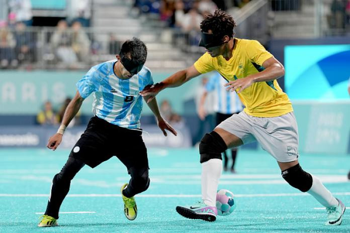 A man in a yellow Brazilian football jersey wearing a black eyeshade plays a football with his foot, while a man in a blue and white Argentina football jersey, also wearing an eyeshade, runs towards him, symbolizing the intensity of Argentina games