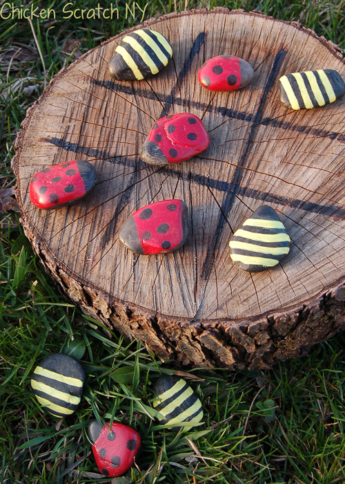 A ladybug and bee-themed tic tac toe game is made from colorfully painted rocks and a wooden stump.