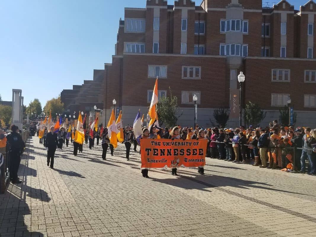 3 color guard members hold a banner that says University of Tennessee, Knoxville, Tennessee while leading the marching band through campus before a Vols home football game