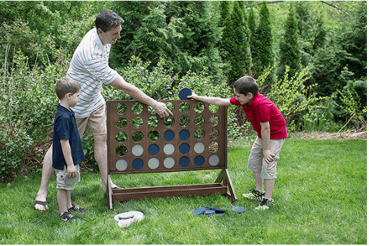 A large wooden DIY Connect Four is set up in a grassy back yard. A father and two young boys are playing with the white and blue game pieces. 