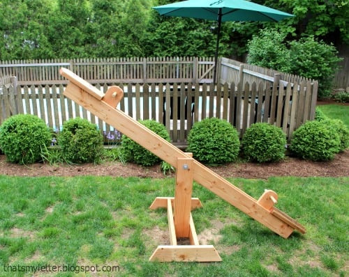 A backyard has a large wooden seesaw displayed on the green grass in front of a fence. 