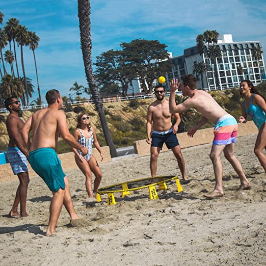 Several people in swimming suits are planning a black and yellow spikeball game on a sandy beach. 