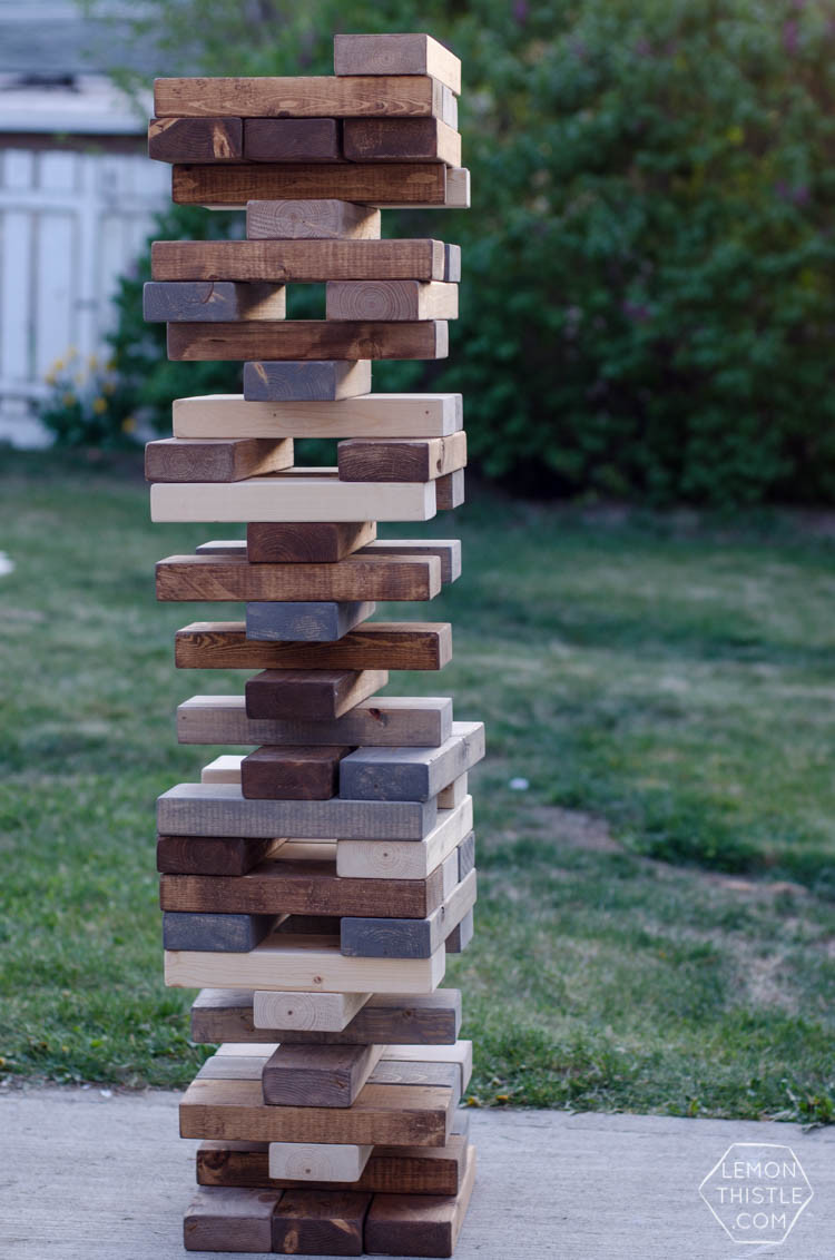 A stack of large wooden Jenga-style blocks are piled high in a backyard with grass behind them. 